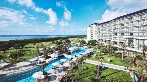 Beachfront resort with a pool, palm trees, and a large hotel building under a bright blue sky