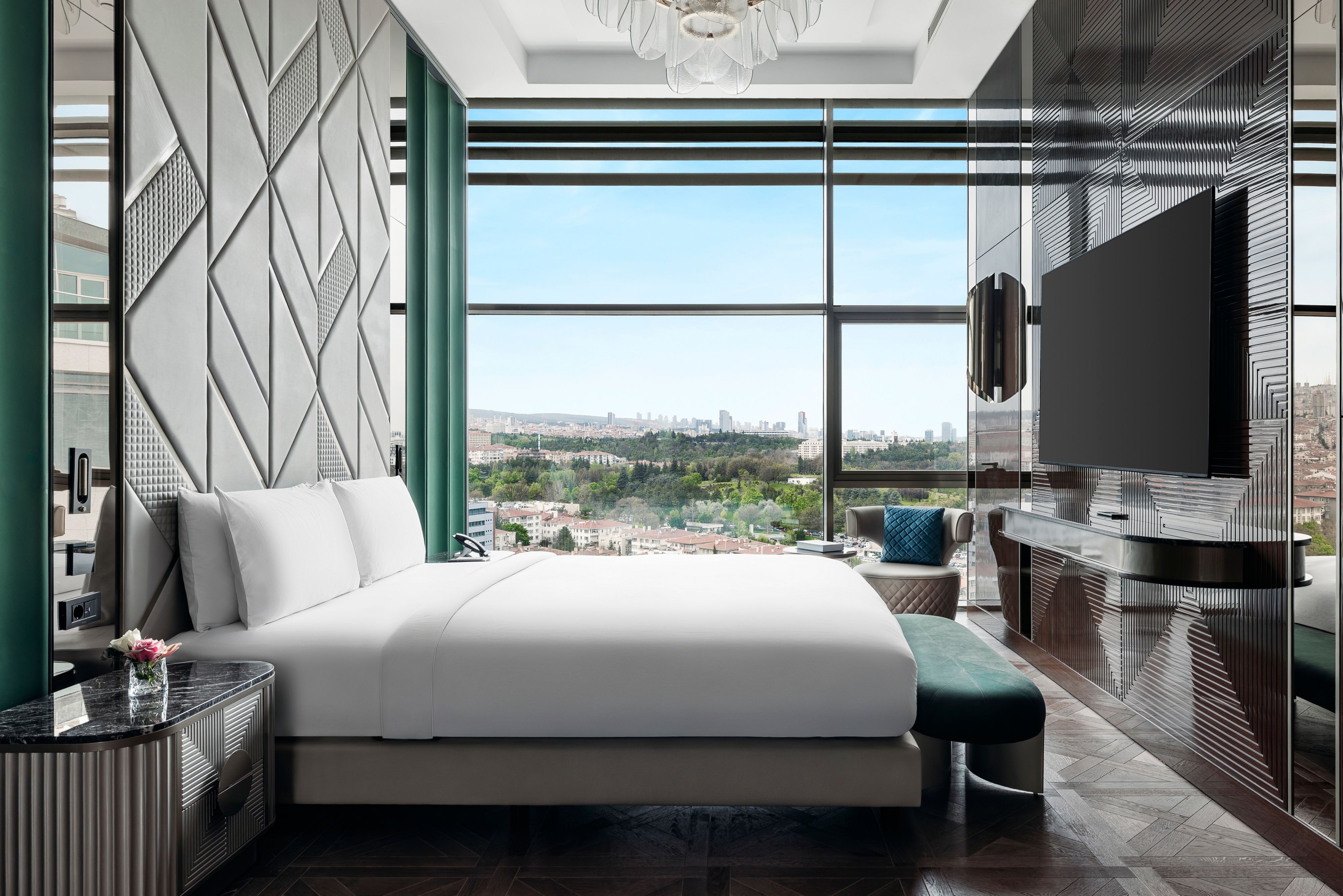Modern hotel bedroom with a geometric feature wall, large window overlooking a city, and a wall-mounted TV.