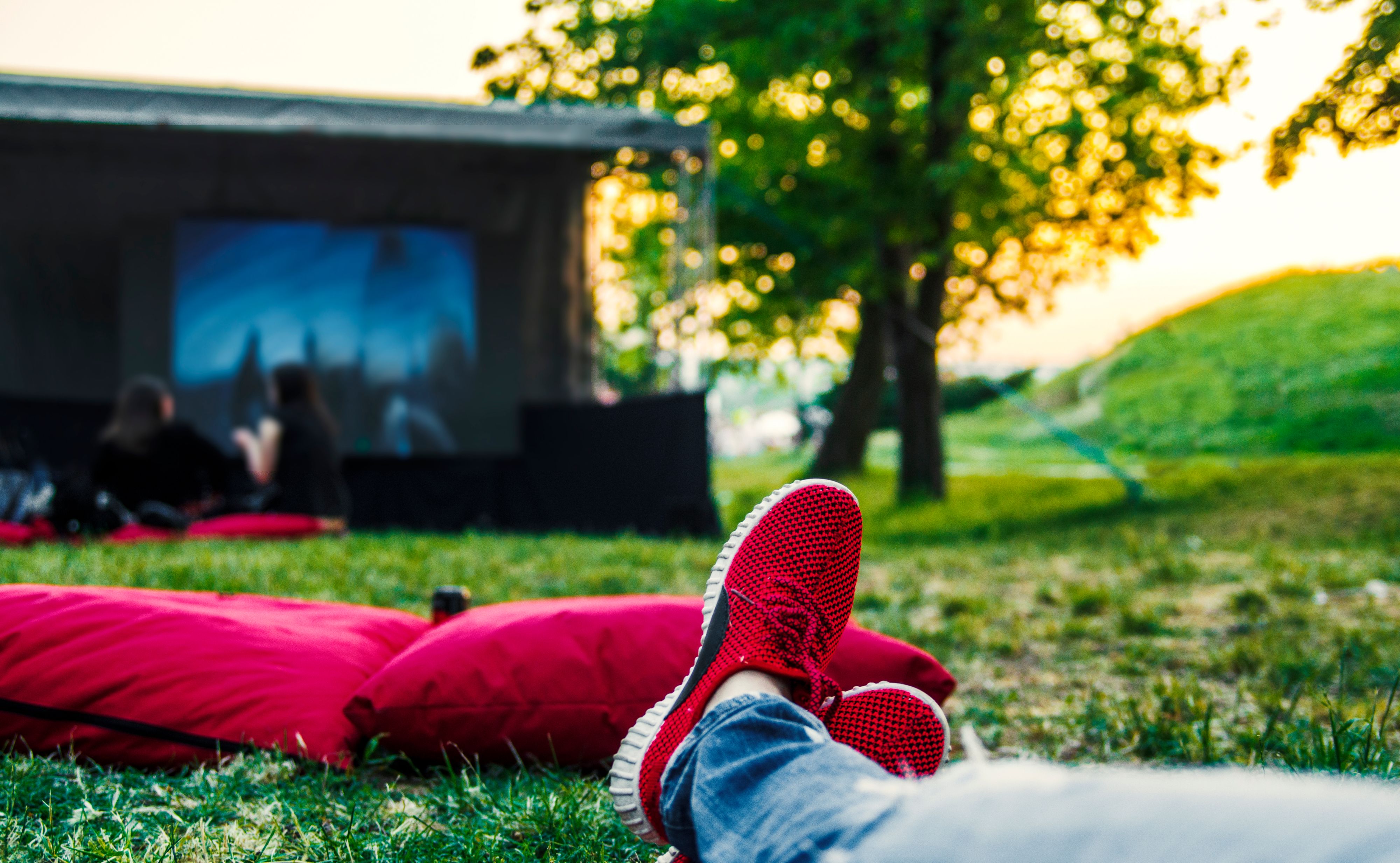Red sneaker shoes on ground,pillows and big screen film projection.Watching movie in open cinema in green nature.Enjoying and relaxing on summer weekend.