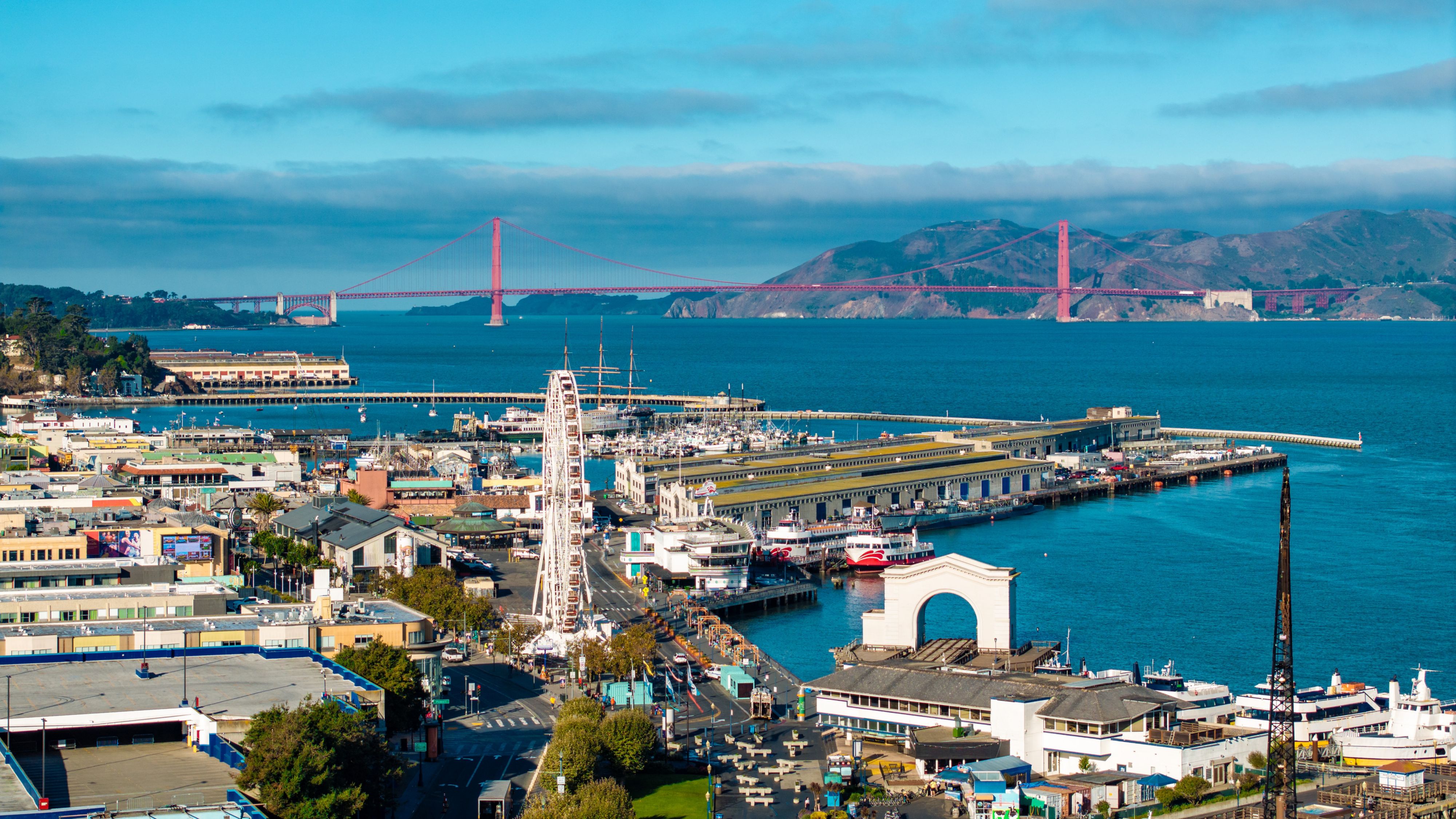 Experience the vibrant aerial view of San Franciscoâ  s Fishermanâ  s Wharf, featuring bustling piers, historic ships, and the iconic Golden Gate Bridge in the background. This famous waterfront captures the energy of the Bay Area and coastal city life.