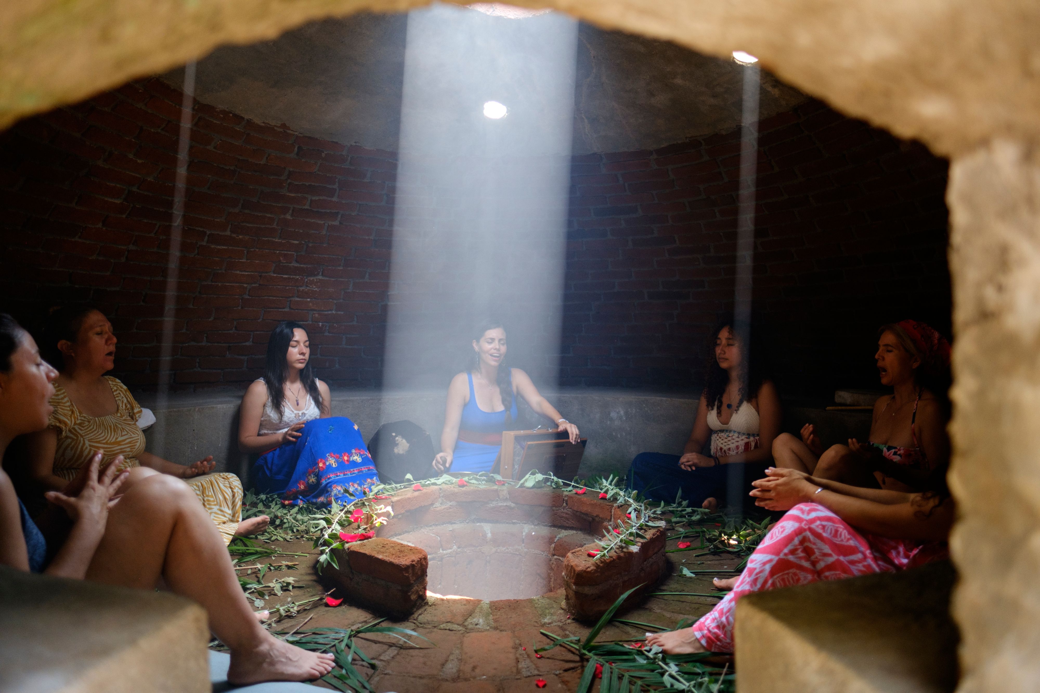 A group of women sitting inside a temazcal with their eyes closed, singing as they are guided by a female guide who plays a harmonium