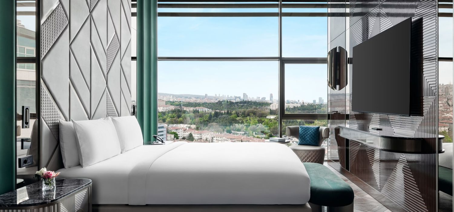 Modern hotel bedroom with a geometric feature wall, large window overlooking a city, and a wall-mounted TV.