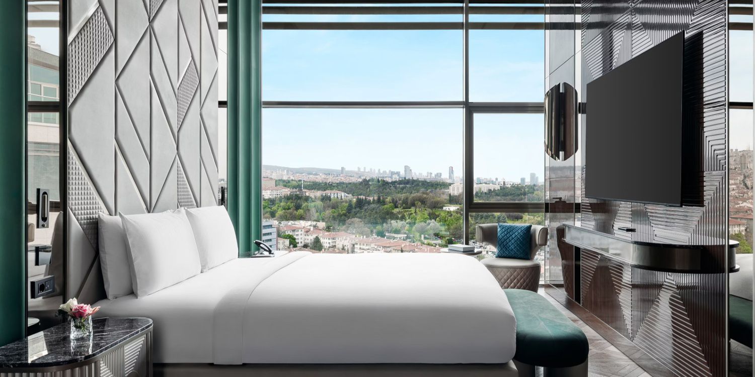 Modern hotel bedroom with a geometric feature wall, large window overlooking a city, and a wall-mounted TV.