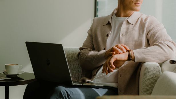 Man sitting with a laptop, checking his watch in a modern lounge chair.