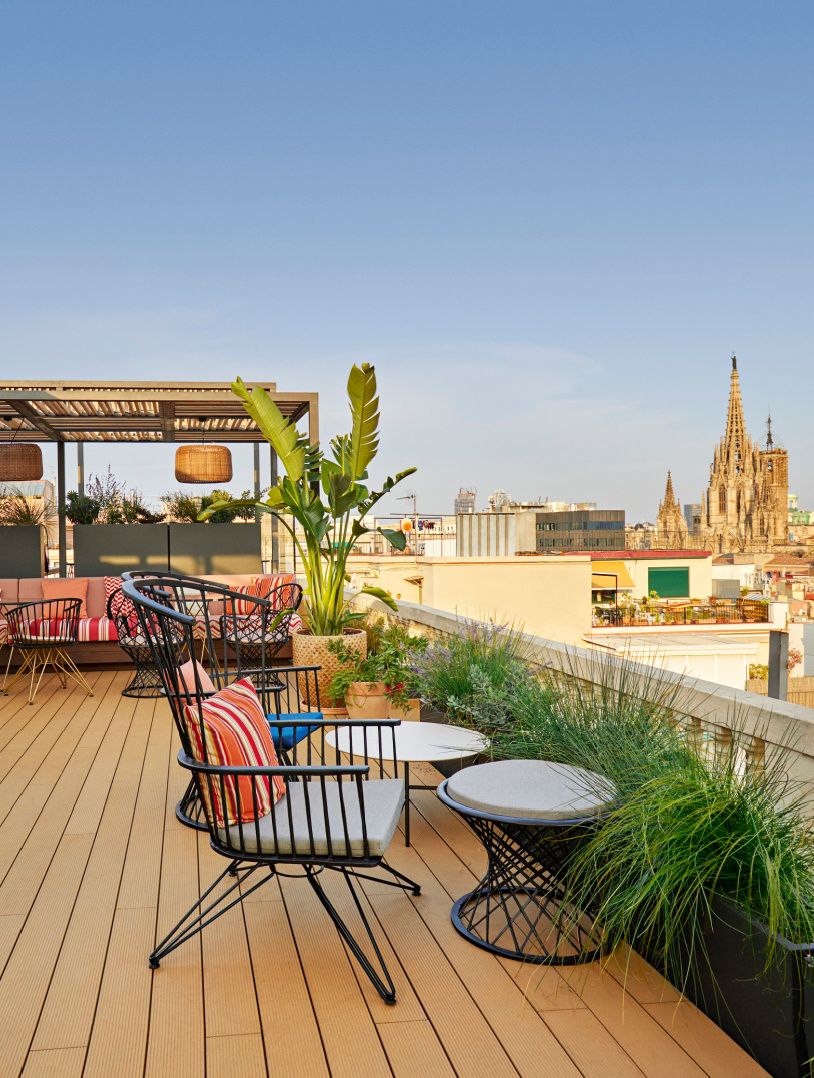 Bar en la azotea con sillones acogedores, plantas exuberantes y vistas de Barcelona.