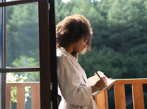 Person in a robe writing in a notebook on a balcony surrounded by greenery