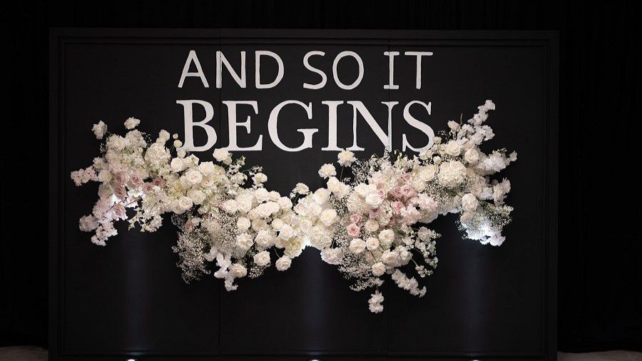 And so it begins sign on wall and flower arrangement