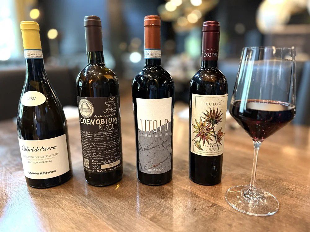 Four bottles of Italian wine displayed beside a glass of red wine on a wooden table.