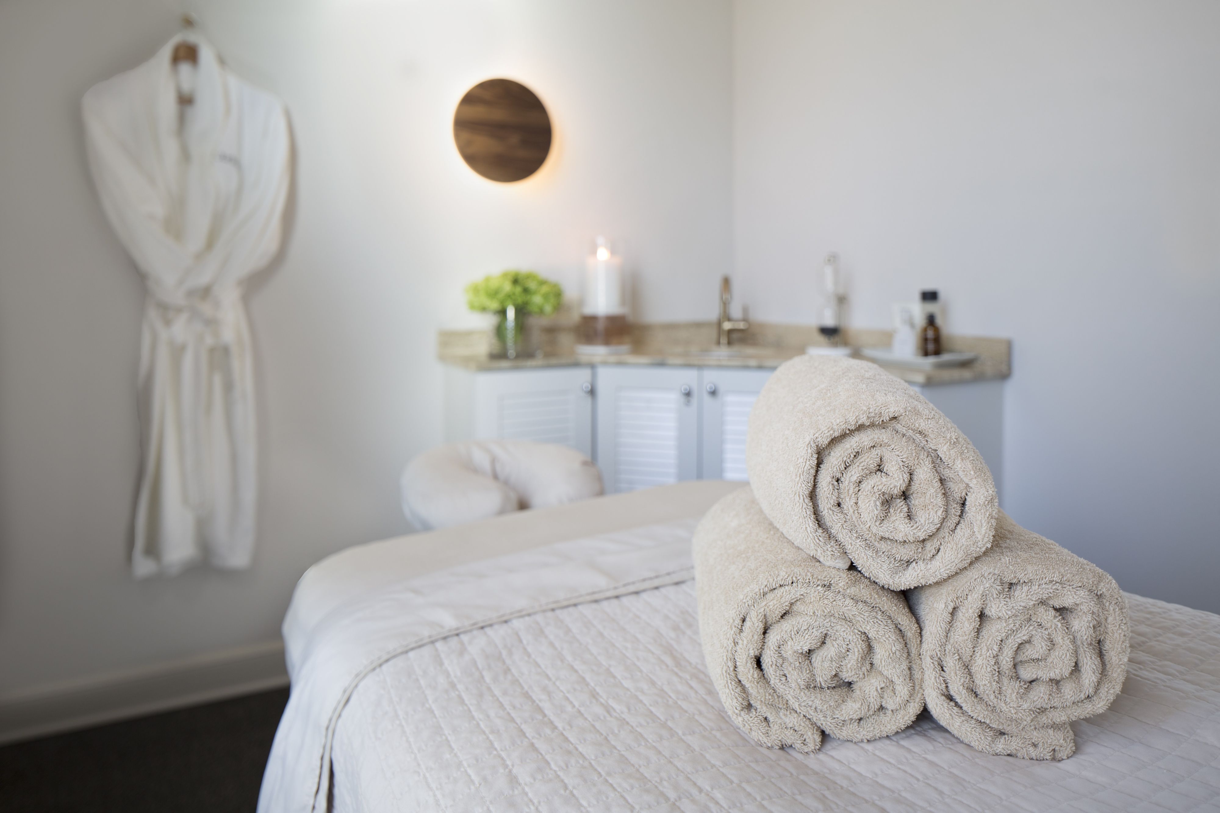 Spa treatment room with a massage table, rolled towels, and a hanging robe.