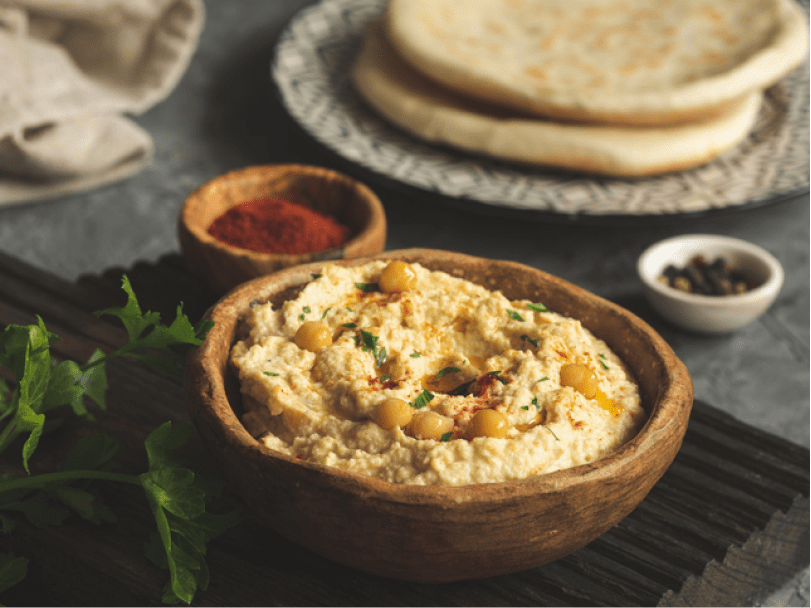 A clay bowl holds fresh hummus. A plate of flatbread and little saucers filled with spices sit behind it.
