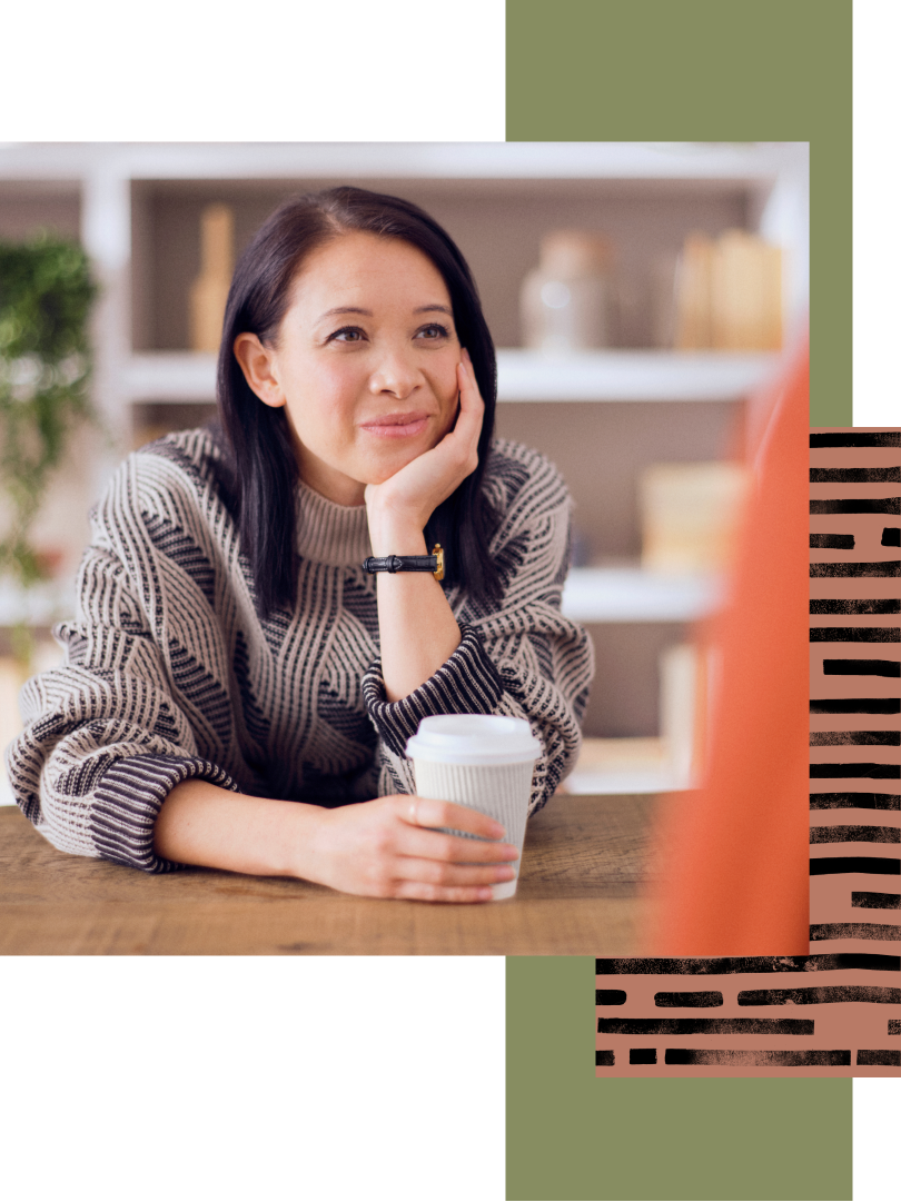An Asian woman wearing a sweater with a stylish, modern print holds a cup of coffee and listens to an unseen companion.