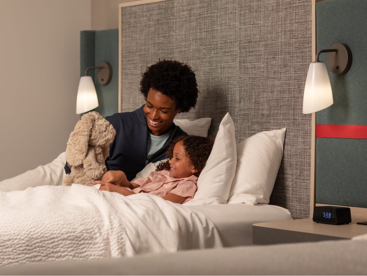 Mère bordant sa fille dans un lit d'hôtel douillet avec un animal en peluche