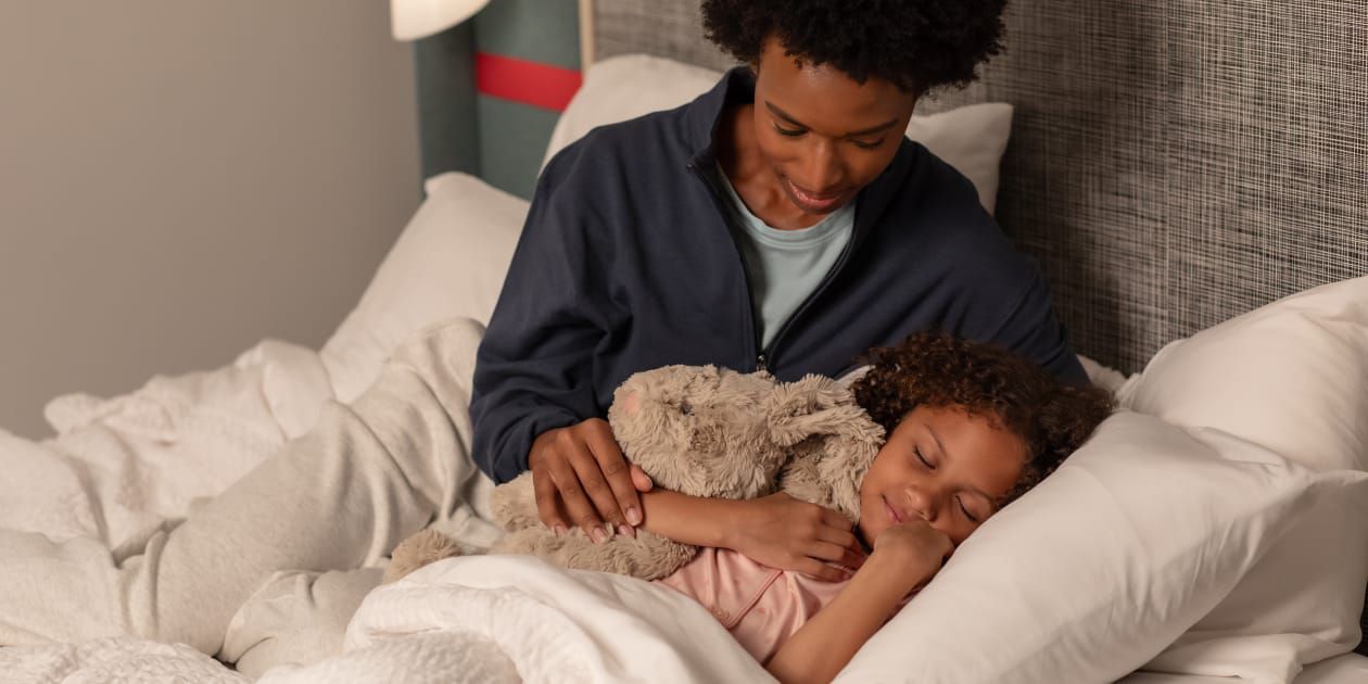 Mom tucking daughter into cozy hotel bed with stuffed animal