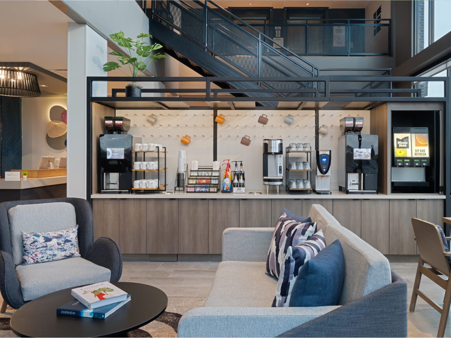 현대적인 호텔 로비에 회색 소파와 안락의자가 놓인 아늑한 휴식 공간이 있습니다. 뒤로는 셀프서비스 커피 바에 머신과 컵이 전시되어 있습니다.