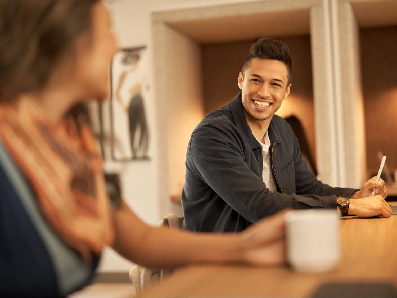 Man smiling while sitting at a table talking to a woman with a coffee mug