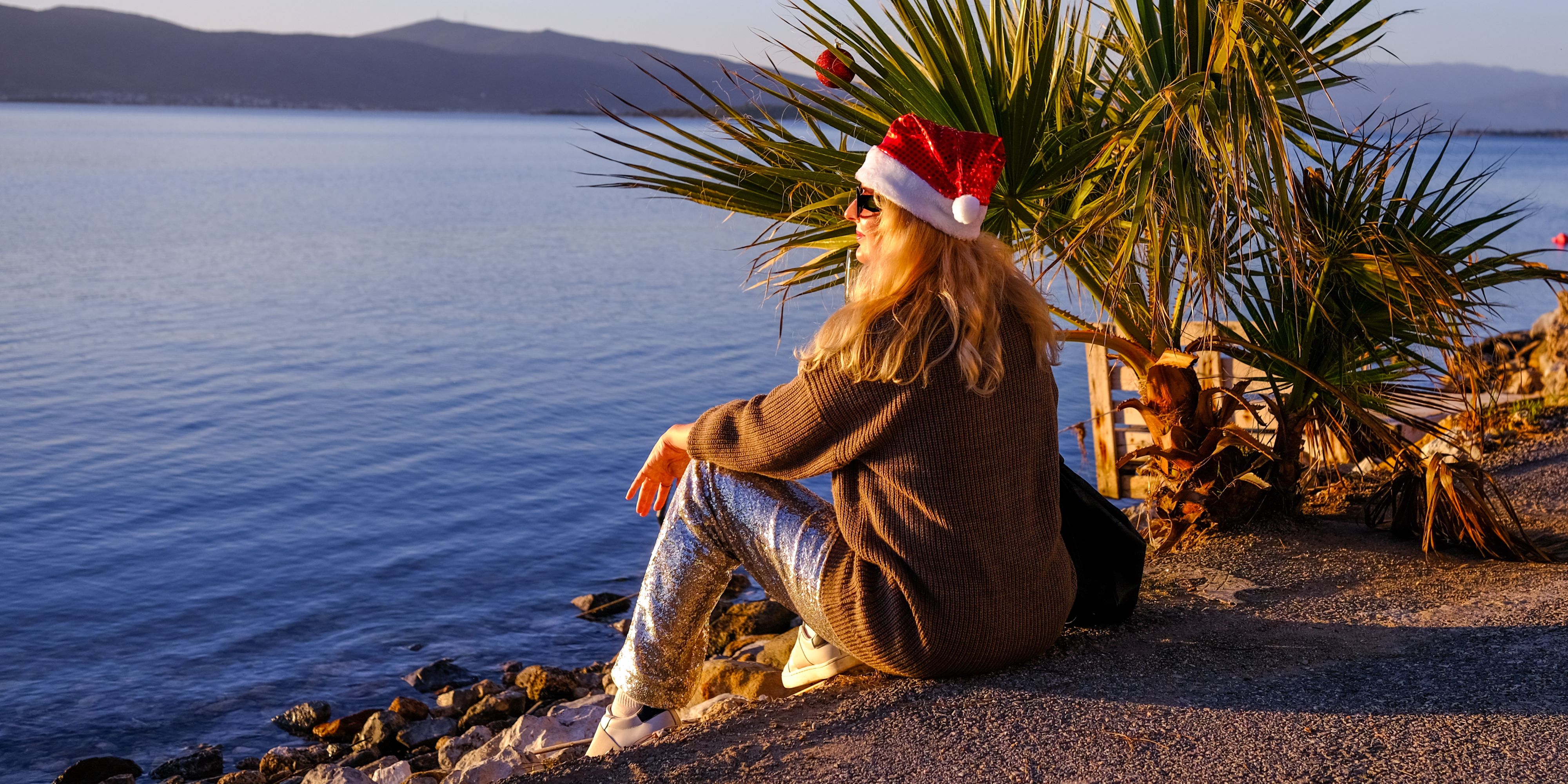 Person wearing a red Santa hat and sunglasses sits on a rocky shoreline beside palm trees, overlooking calm blue water and distant mountains in warm golden-hour light.