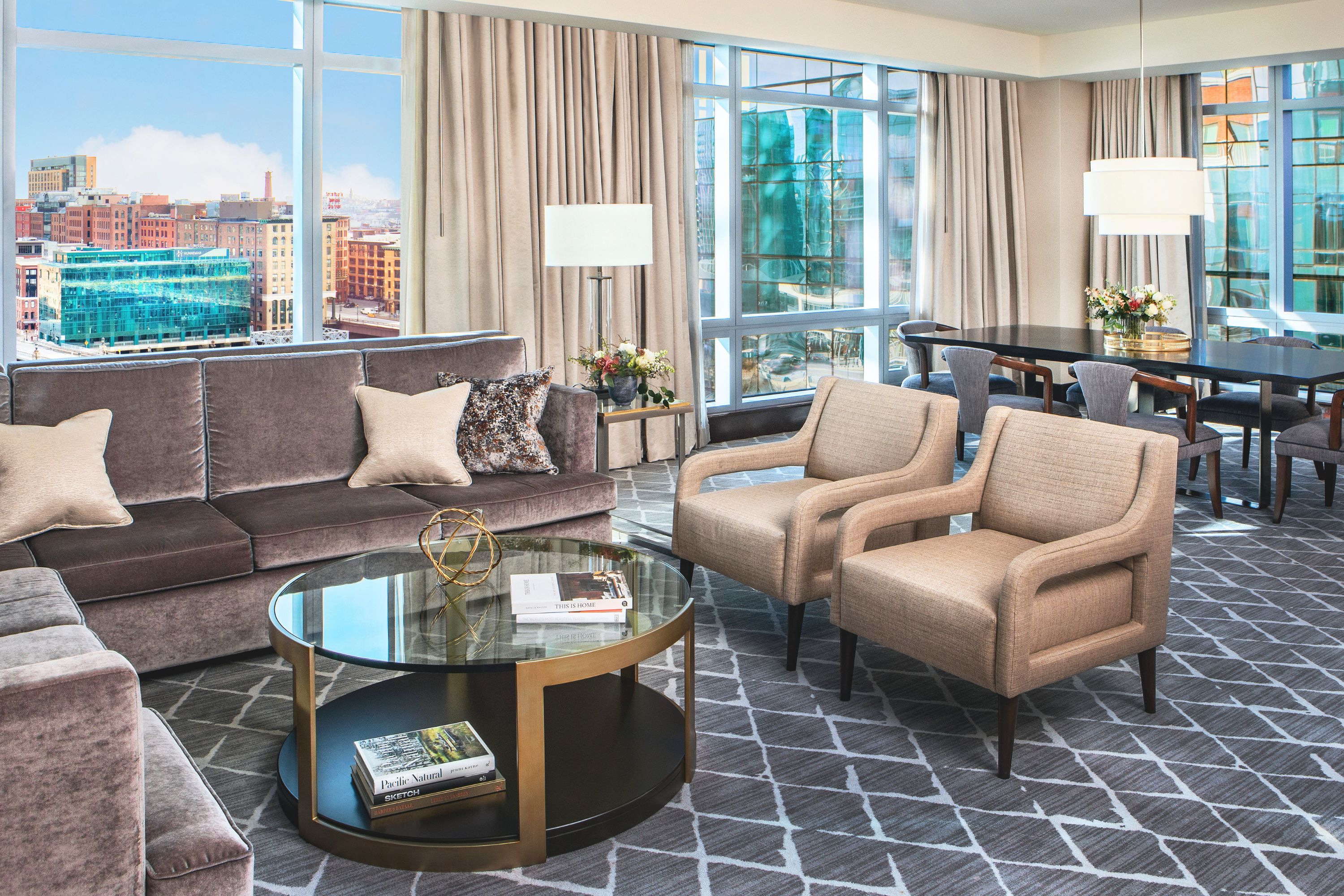 Spacious living and dining area with sofa, armchairs, round coffee table, and dining table set near large windows overlooking the city.