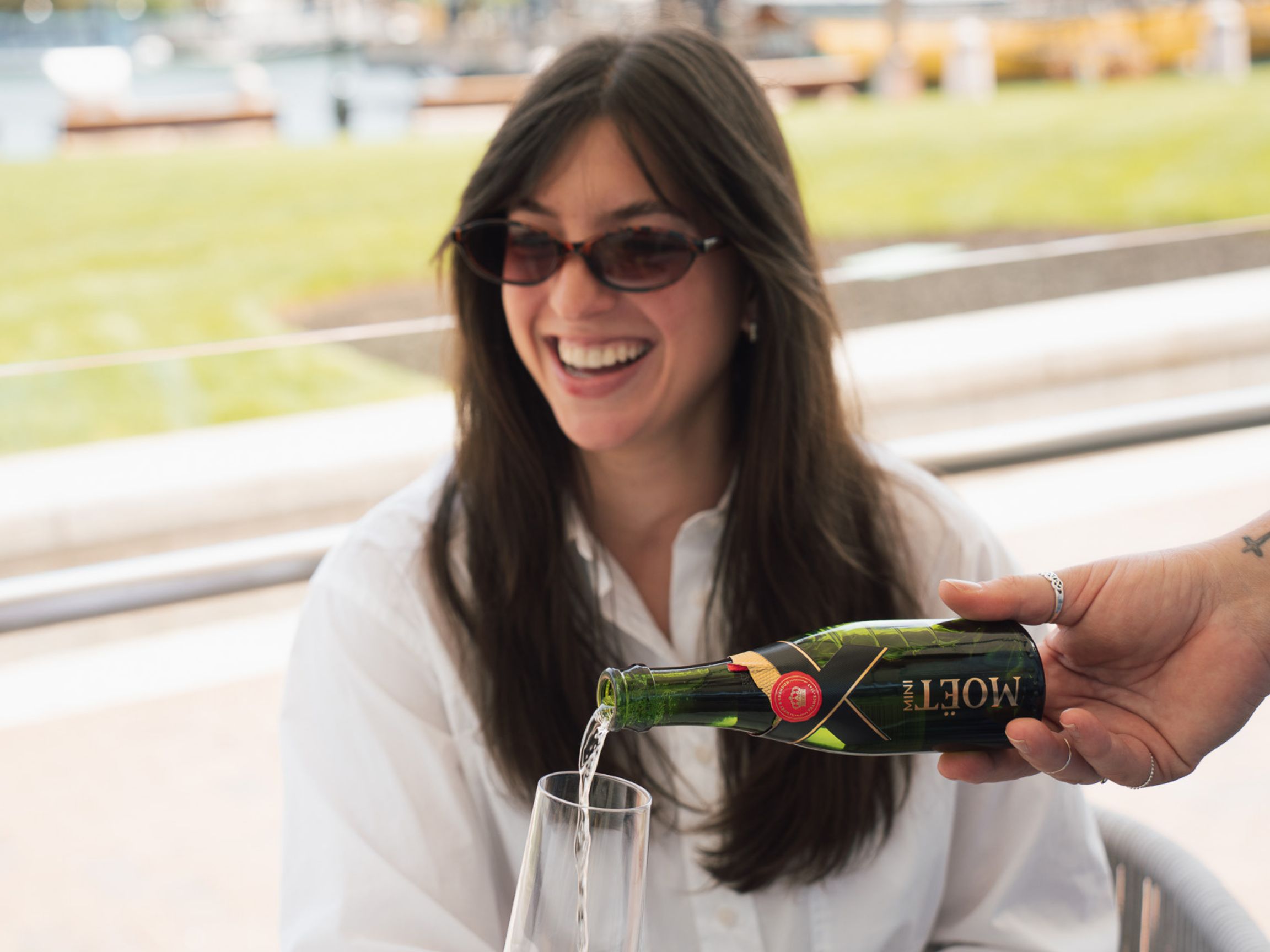 Guest seated on an outdoor terrace as sparkling wine is poured into a flute, with waterfront views softly blurred in the background.