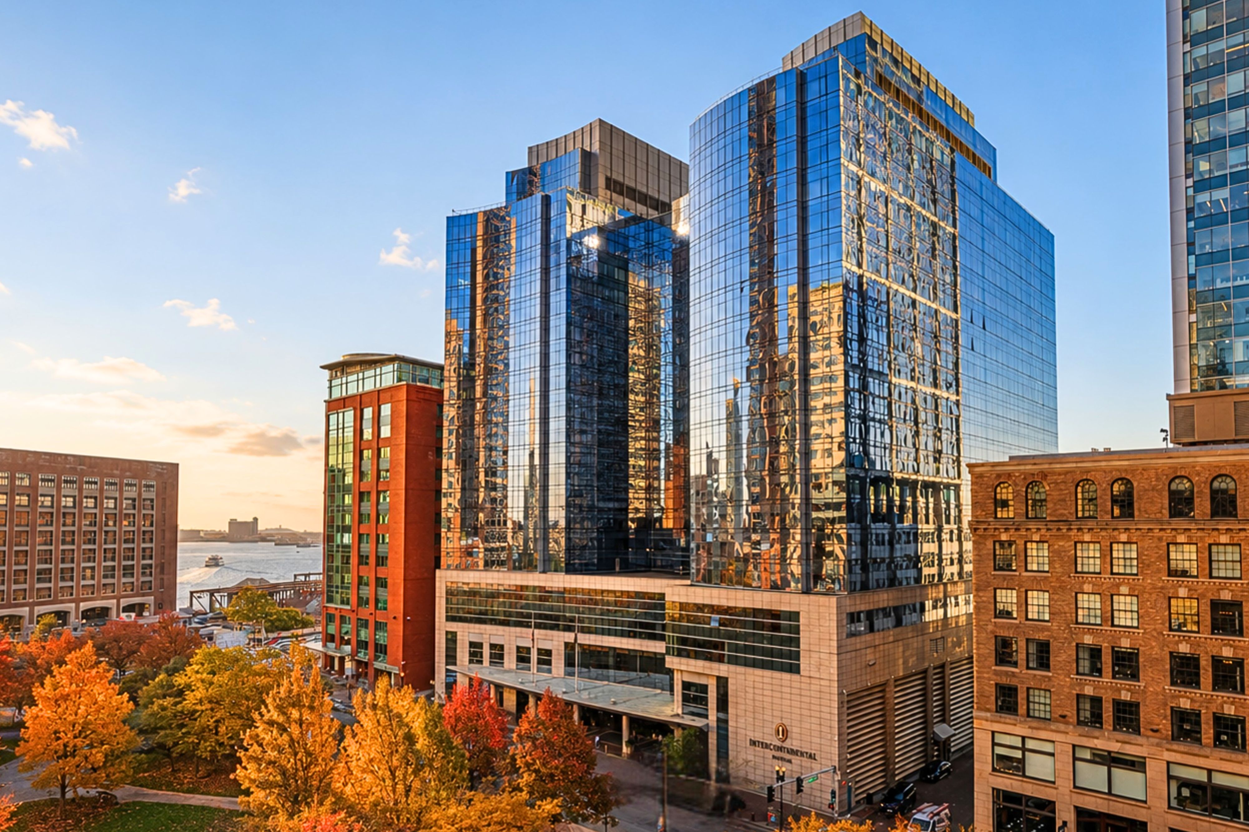 InterContinental Boston exterior at sunset, featuring a modern glass facade reflecting the city skyline, with colorful autumn trees and Boston Harbor visible nearby.