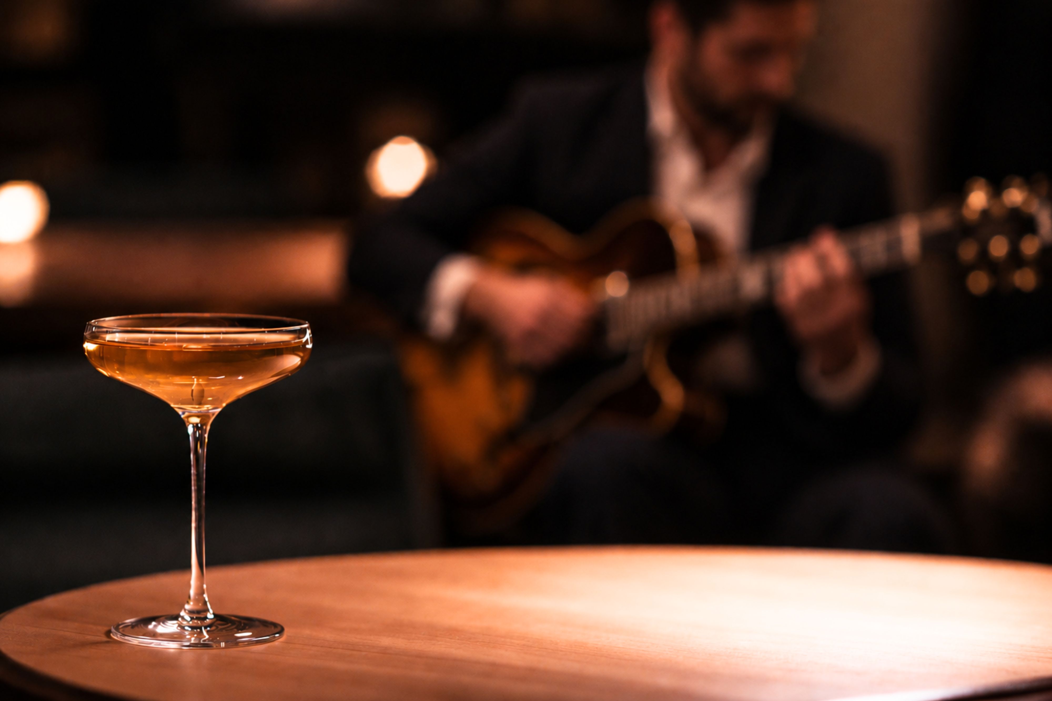 Cocktail in a coupe glass on a lounge table, with a guitarist performing in the softly lit background, creating an intimate atmosphere for live music and evening drinks.