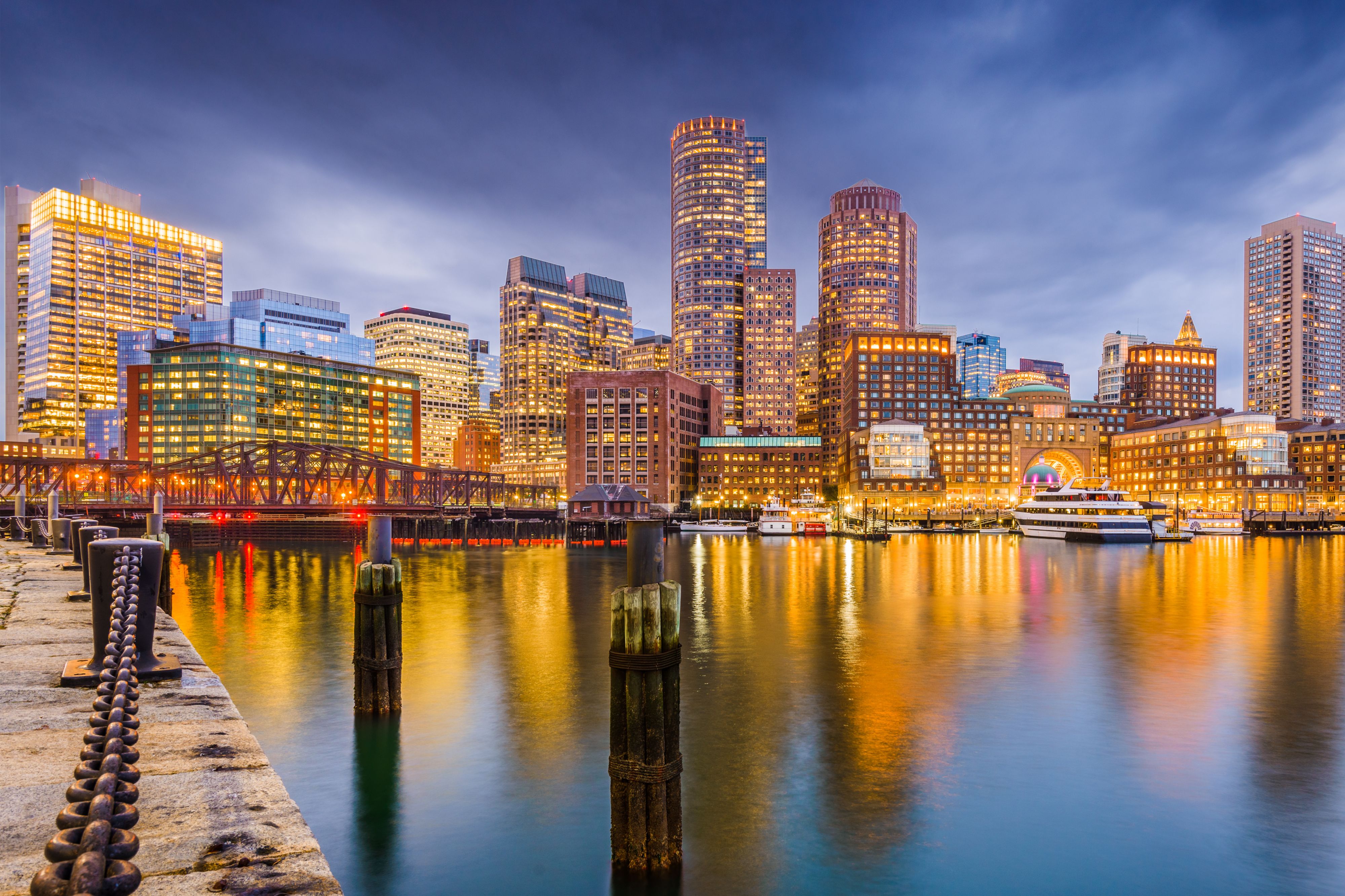 Boston Harbor waterfront at dusk with illuminated downtown skyscrapers, a pedestrian bridge, and boats along the harbor, reflected in the calm water beneath a dramatic evening sky.