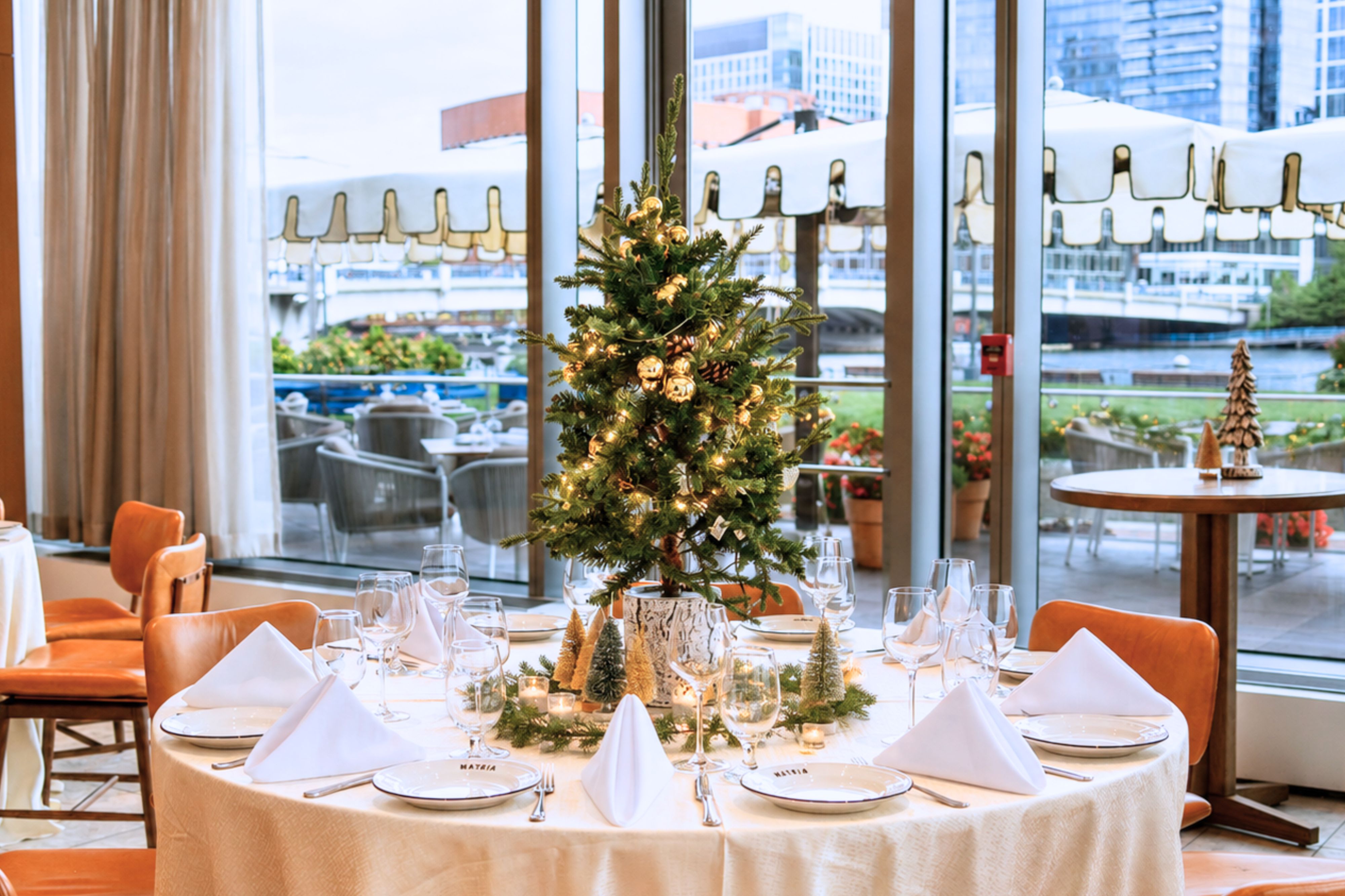 Holiday dining table set for a special gathering, featuring a decorated Christmas tree centerpiece, elegant place settings, candlelight accents, and waterfront views through floor-to-ceiling windows.