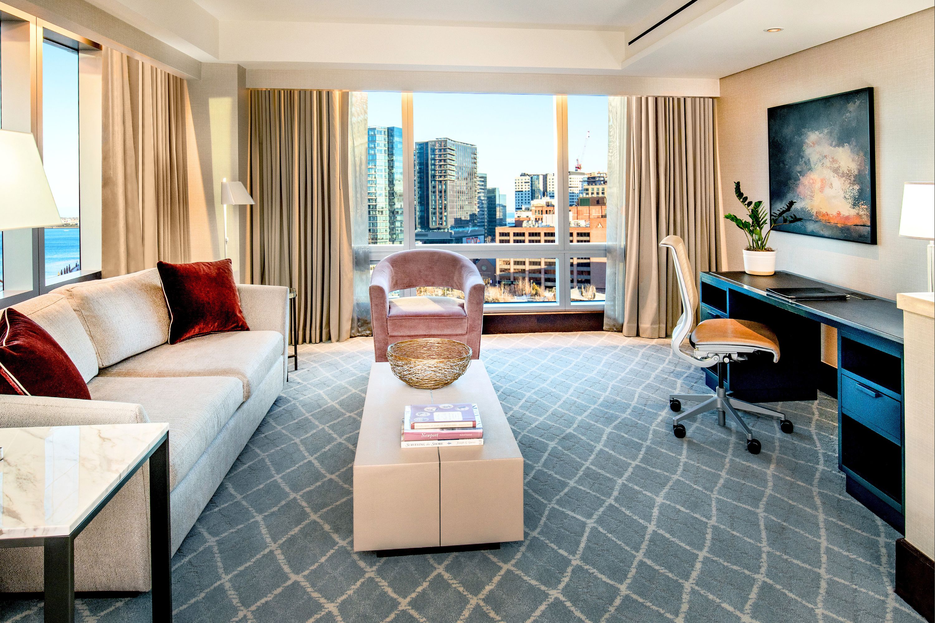 Suite living area with sofa, armchair, coffee table, and work desk, positioned in front of large windows with city views.