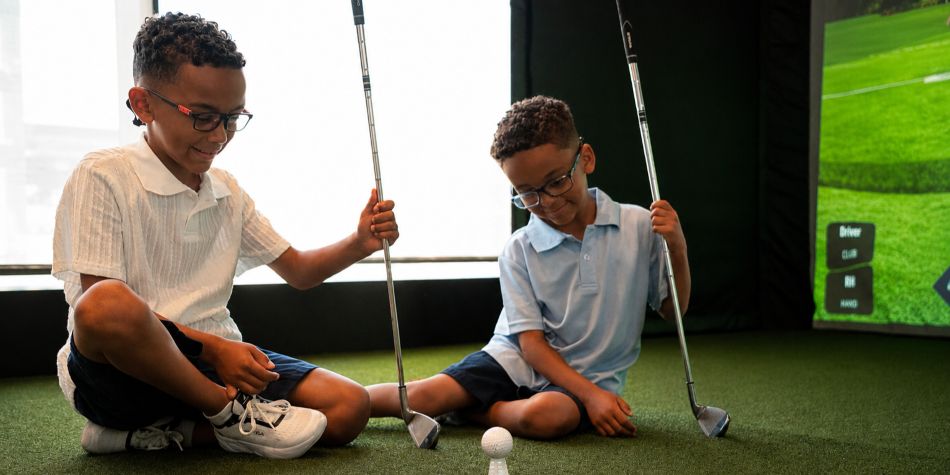 Two kids hold golf clubs on turf in an indoor golf simulator.