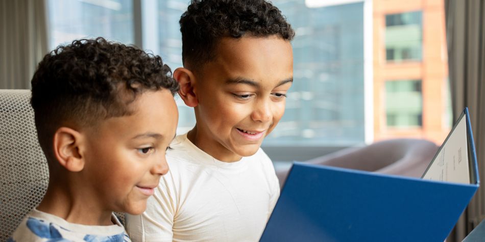Two smiling kids read a blue Kids Club menu by a sunny window.