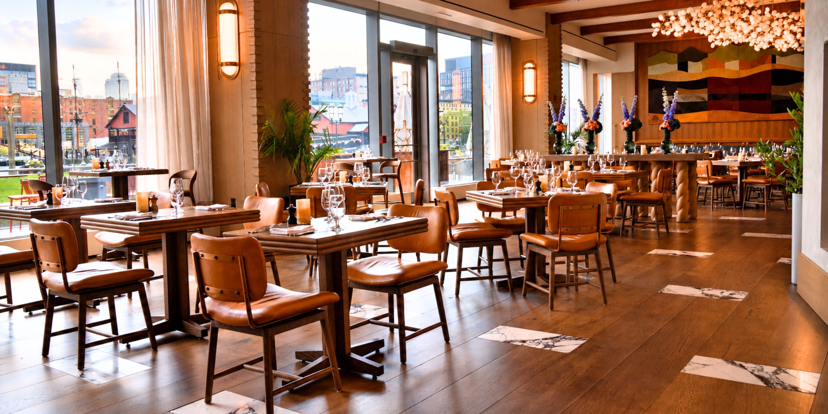 Spacious waterfront restaurant with floor-to-ceiling windows, wood furnishings, set dining tables, and views of the harbor, creating a warm and inviting dining atmosphere.