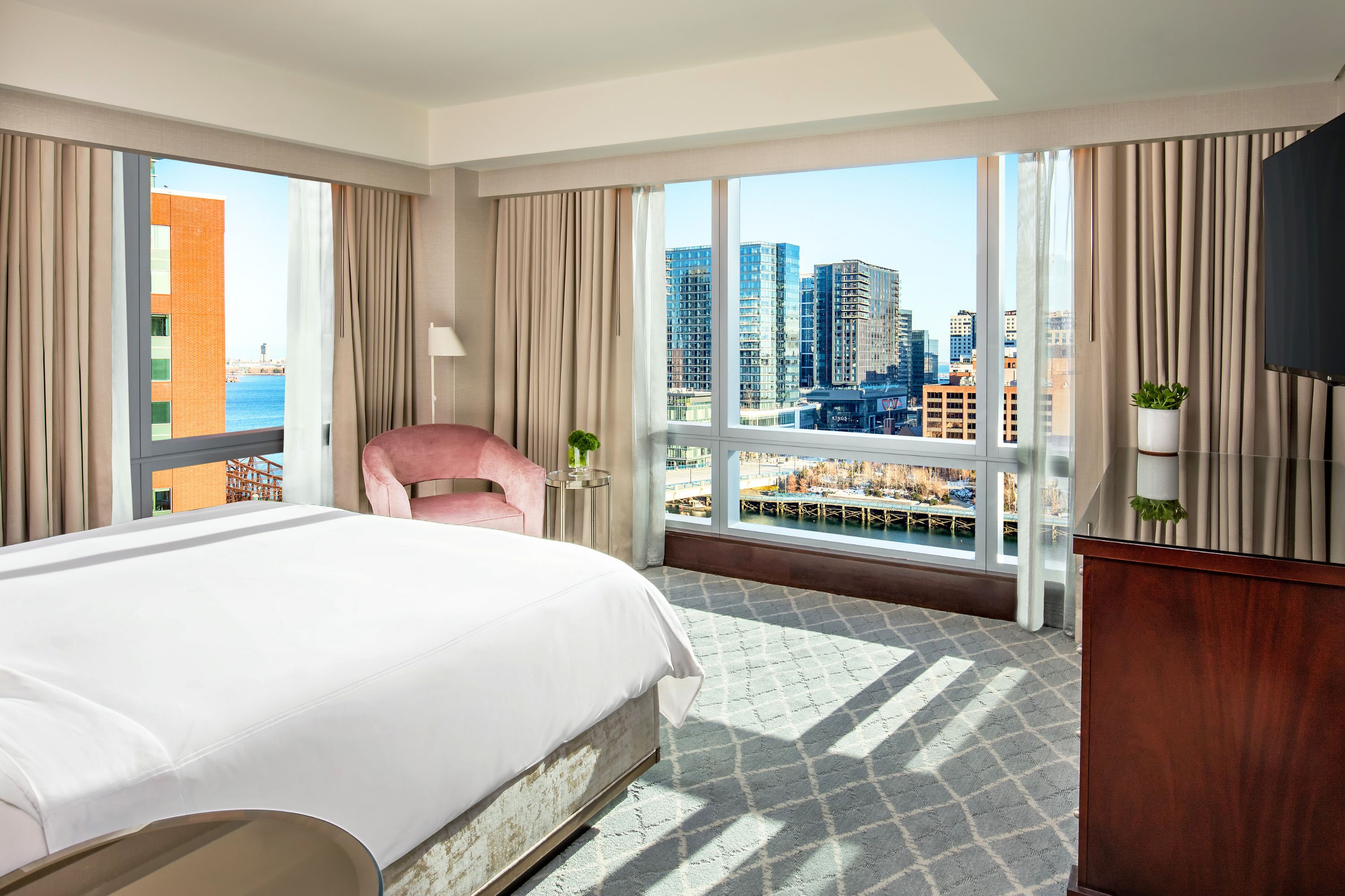 Bedroom with a king bed near expansive corner windows offering wide views of Boston waterfront and surrounding buildings.