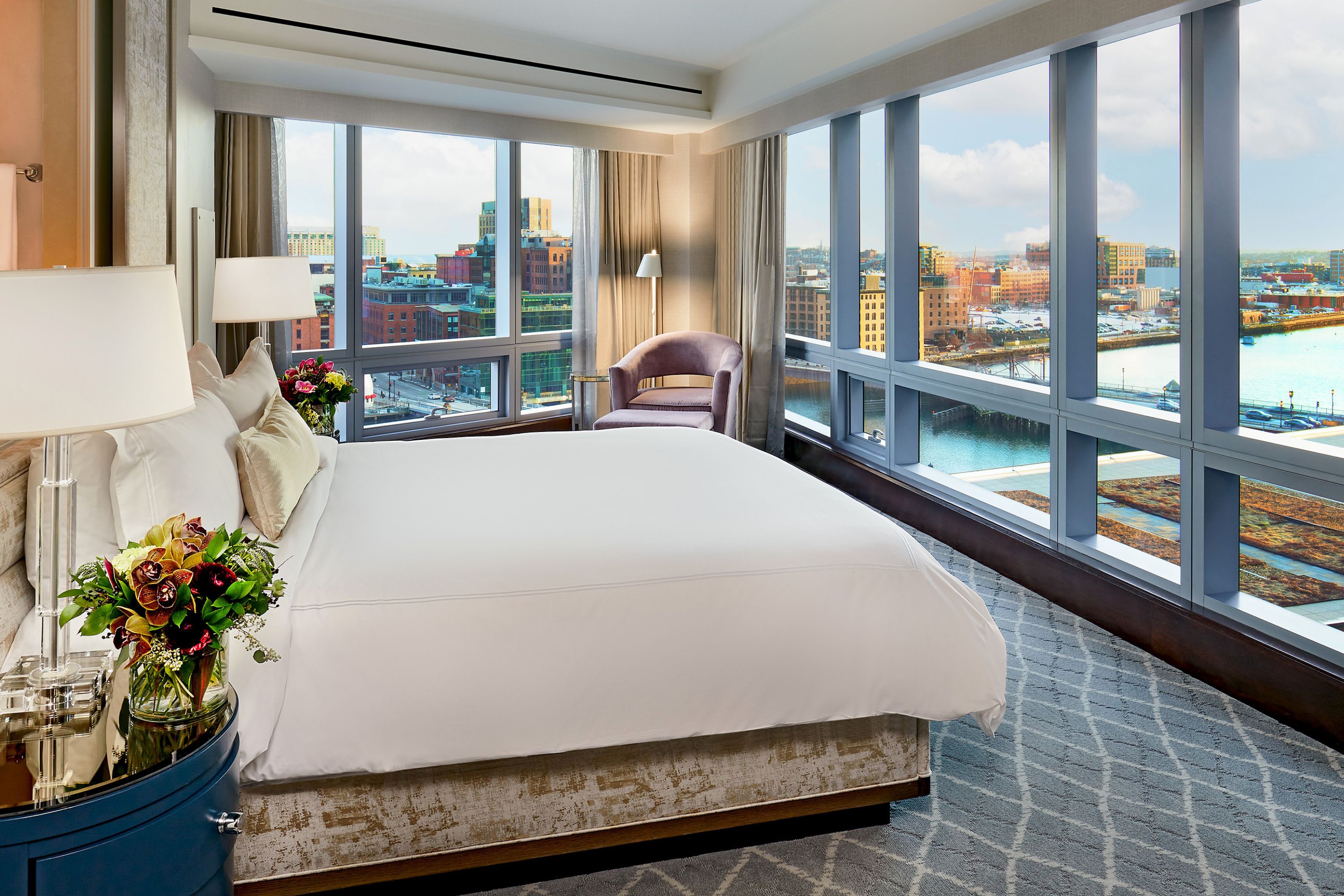 Bedroom with a king bed beside floor-to-ceiling windows showing Boston skyline and waterfront views, with a chair in the corner.