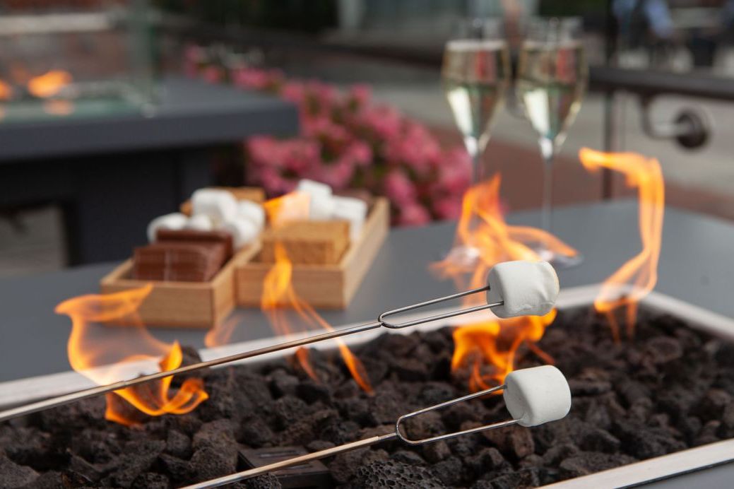 Guests roast marshmallows over an outdoor fire table, with s’mores ingredients and sparkling wine set nearby on a patio at InterContinental Boston.