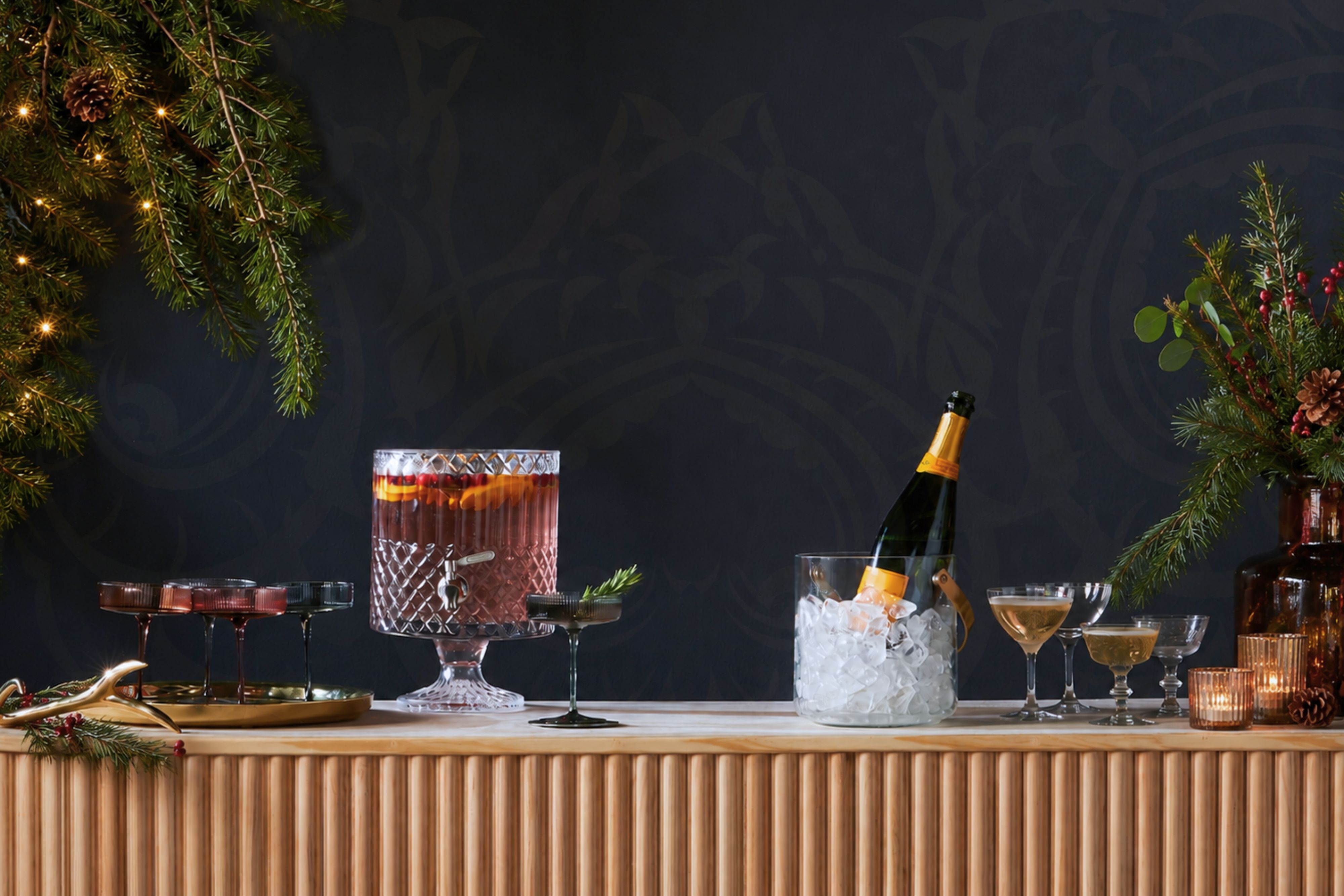 Festive holiday cocktail display featuring a crystal punch bowl, champagne chilling on ice, elegant coupe glasses, evergreen garlands, pinecones, and candlelight on a stylish bar. 