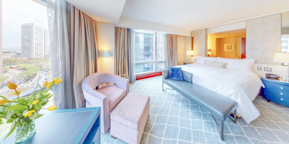 Spacious bedroom with a king bed, upholstered bench, armchair, and floor-to-ceiling windows offering city views.