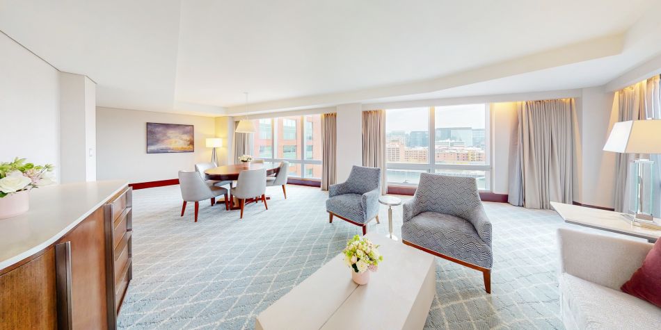 Spacious suite living and dining area with a table for six, two armchairs, and floor-to-ceiling windows showing city views.