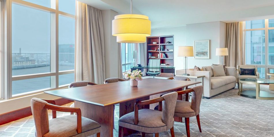 Wood dining table with cushioned chairs set near large windows, with a living area and sofa visible in the background.