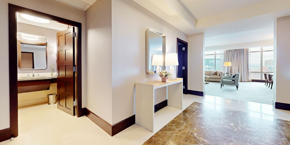 Suite entry hallway with console table and mirror leading into a bright living area with seating and large windows.