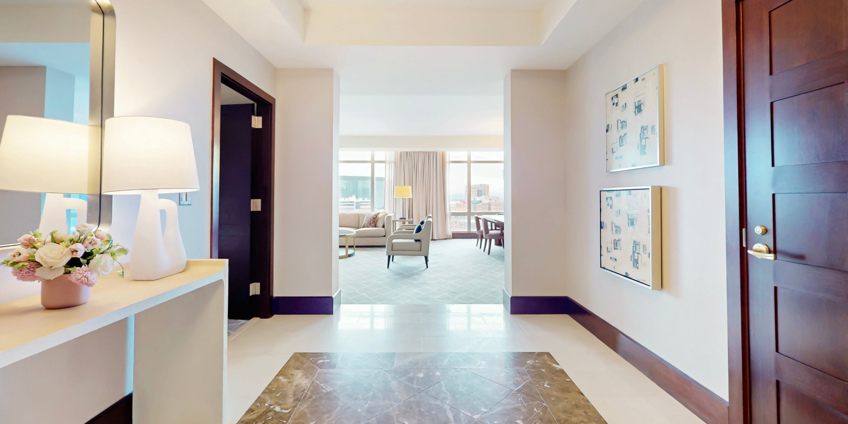 Spacious suite entry with console table, lamp, and artwork leading to a bright living area with sofa, armchair, dining table, and large windows with city views.