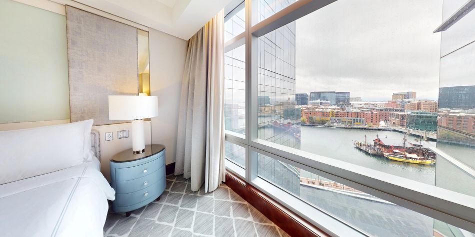 Bedroom corner with a bedside table and lamp next to large windows overlooking the waterfront and city skyline.