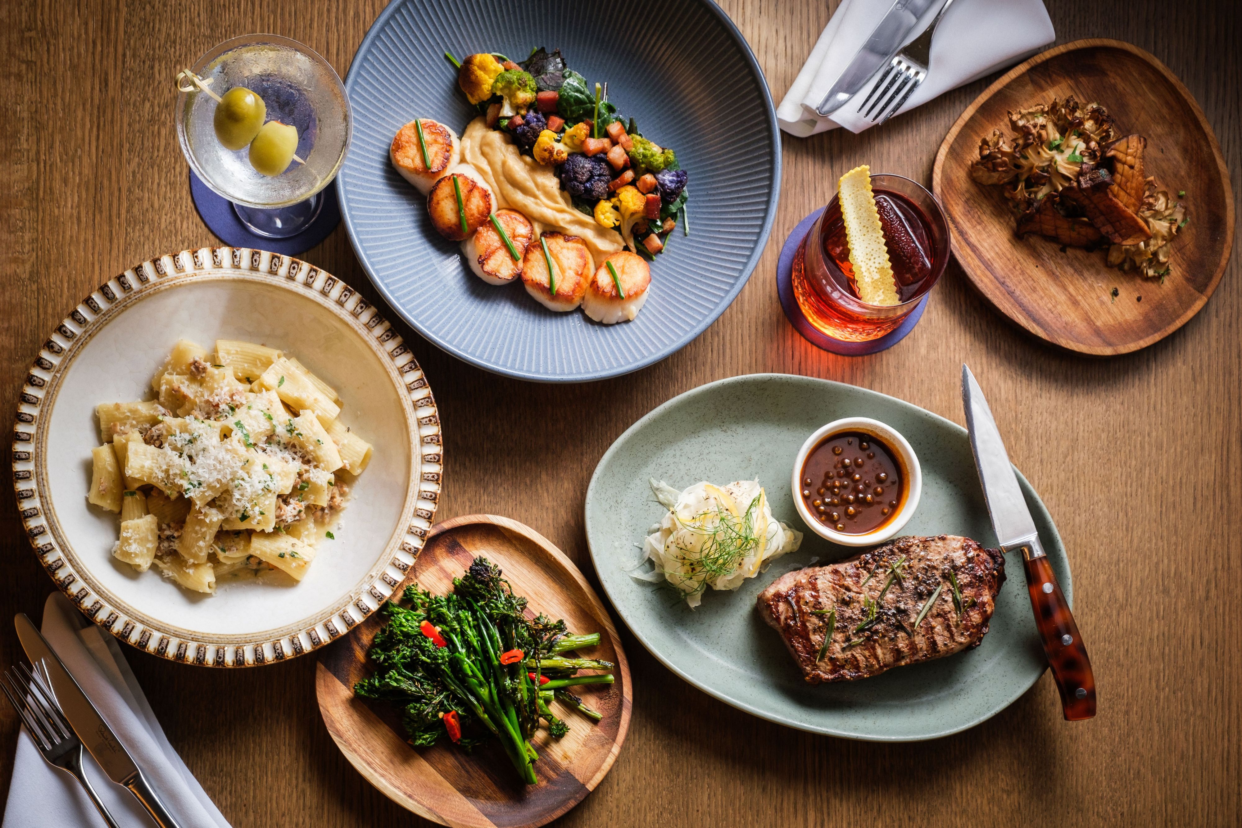 Overhead view of a restaurant table featuring grilled steak, seared scallops with vegetables, rigatoni pasta, broccolini, roasted mushrooms, and two cocktails on a wooden tabletop.
