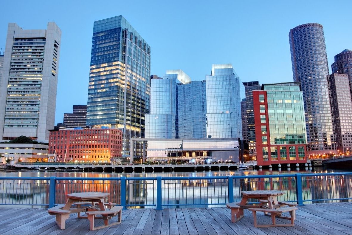Boston Harbor skyline seen from a waterfront deck with picnic tables.