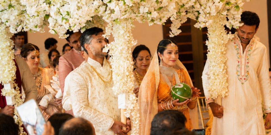 Wedding ceremony processional beneath a canopy of white flowers, surrounded by guests and traditional floral décor in an indoor event space.