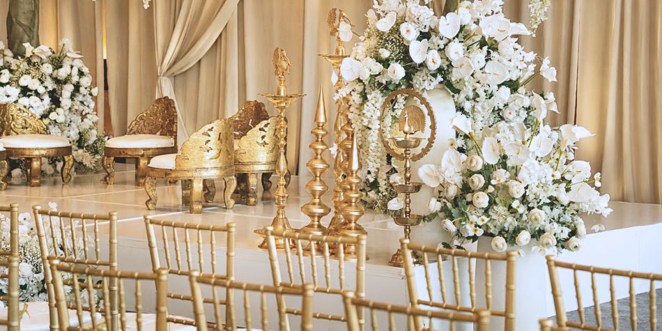 Wedding ceremony stage adorned with white floral arrangements, ornate gold seating, and decorative candle stands in an elegant ballroom setting.