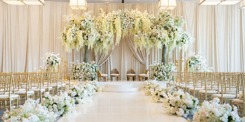 Elegant indoor wedding ceremony venue featuring a flower-lined aisle, gold chairs, and a grand floral backdrop with cascading white blooms and draped curtains.