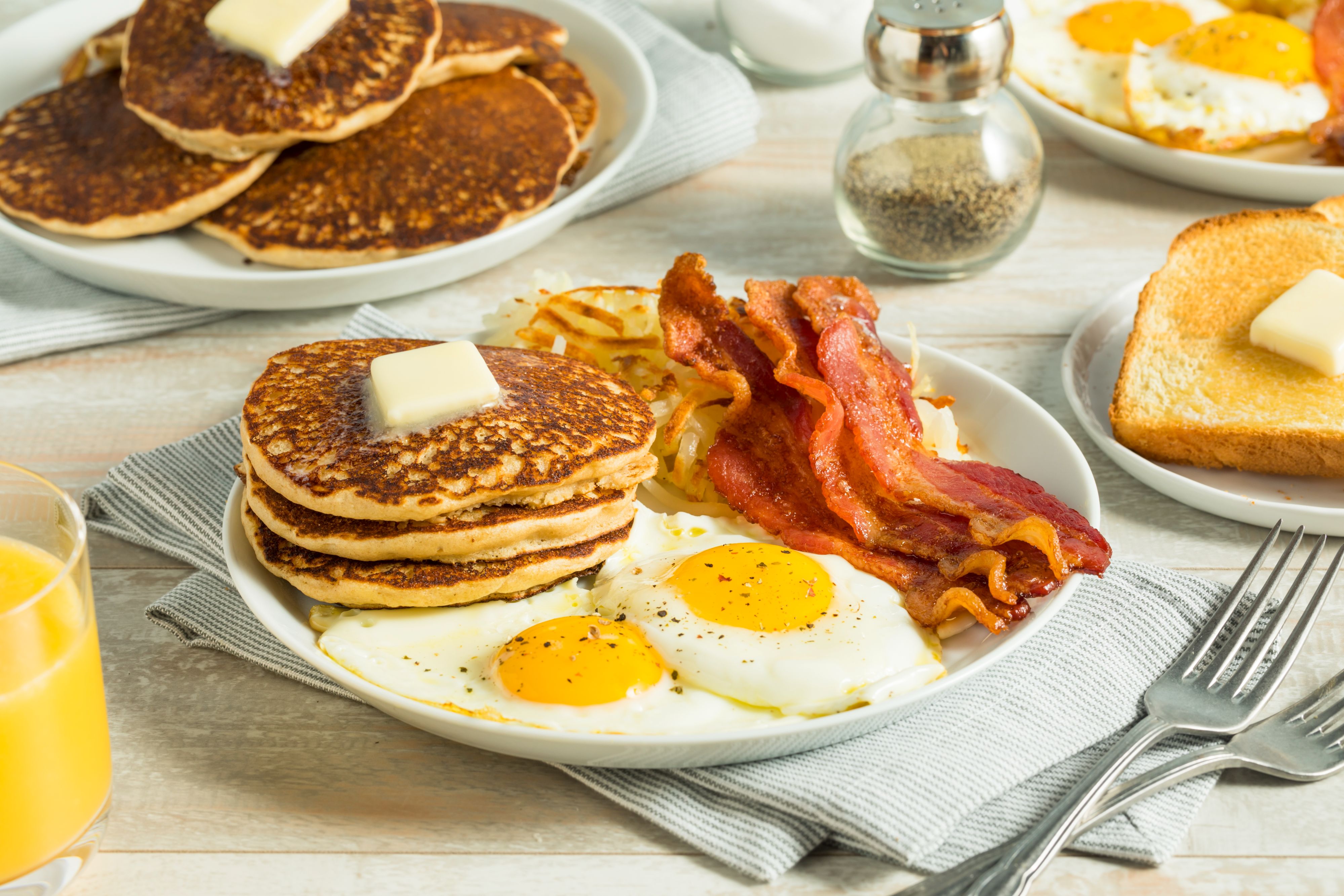 Plate of breakfast food with stacked pancakes topped with butter, two sunny-side-up eggs, crispy bacon, and hash browns, with toast and orange juice nearby.