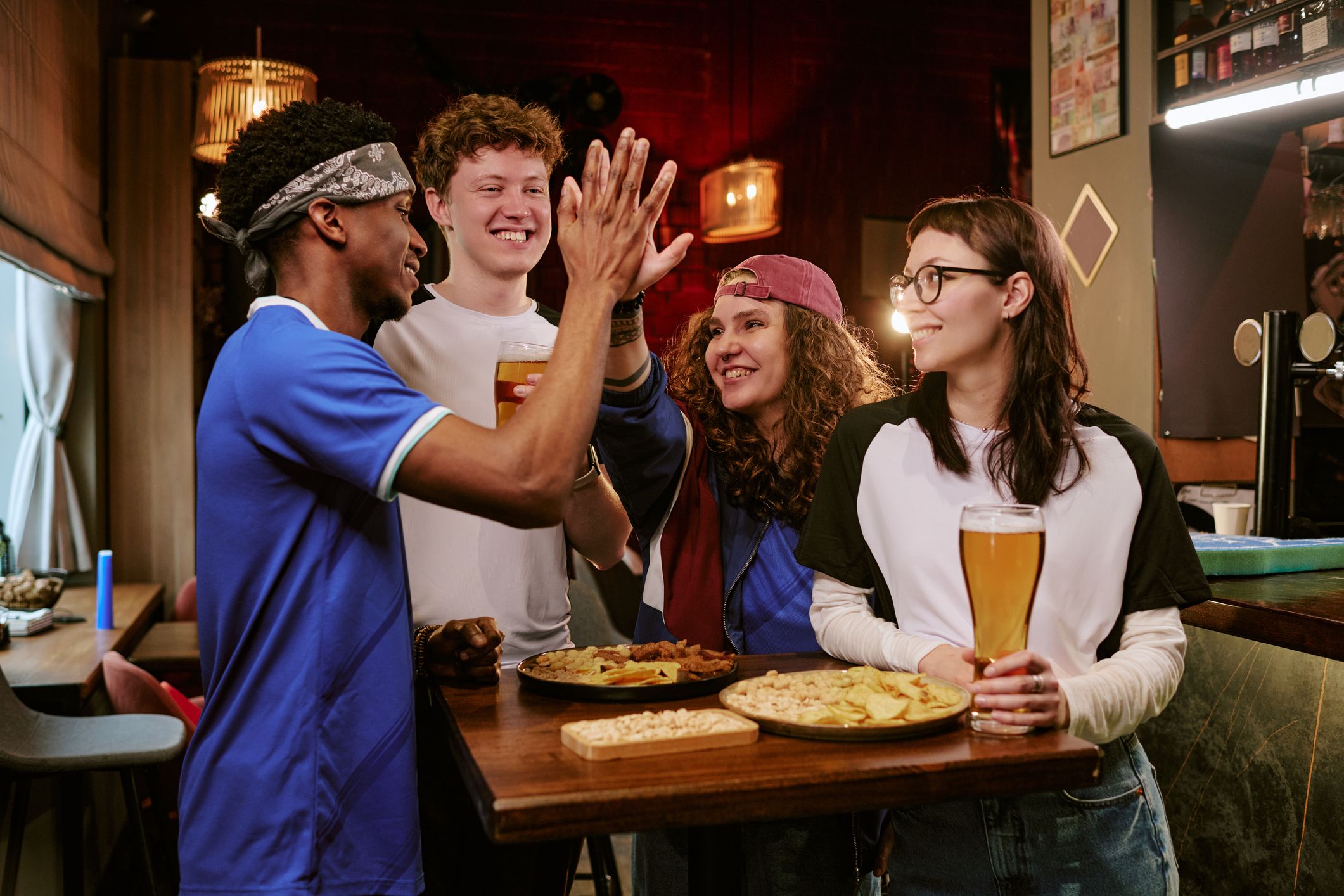 Multiethnic young adults celebrating sports match, high fiving and drinking beer at pub table with snacks. Ideal for fan culture, social gathering, hospitality marketing, medium shot