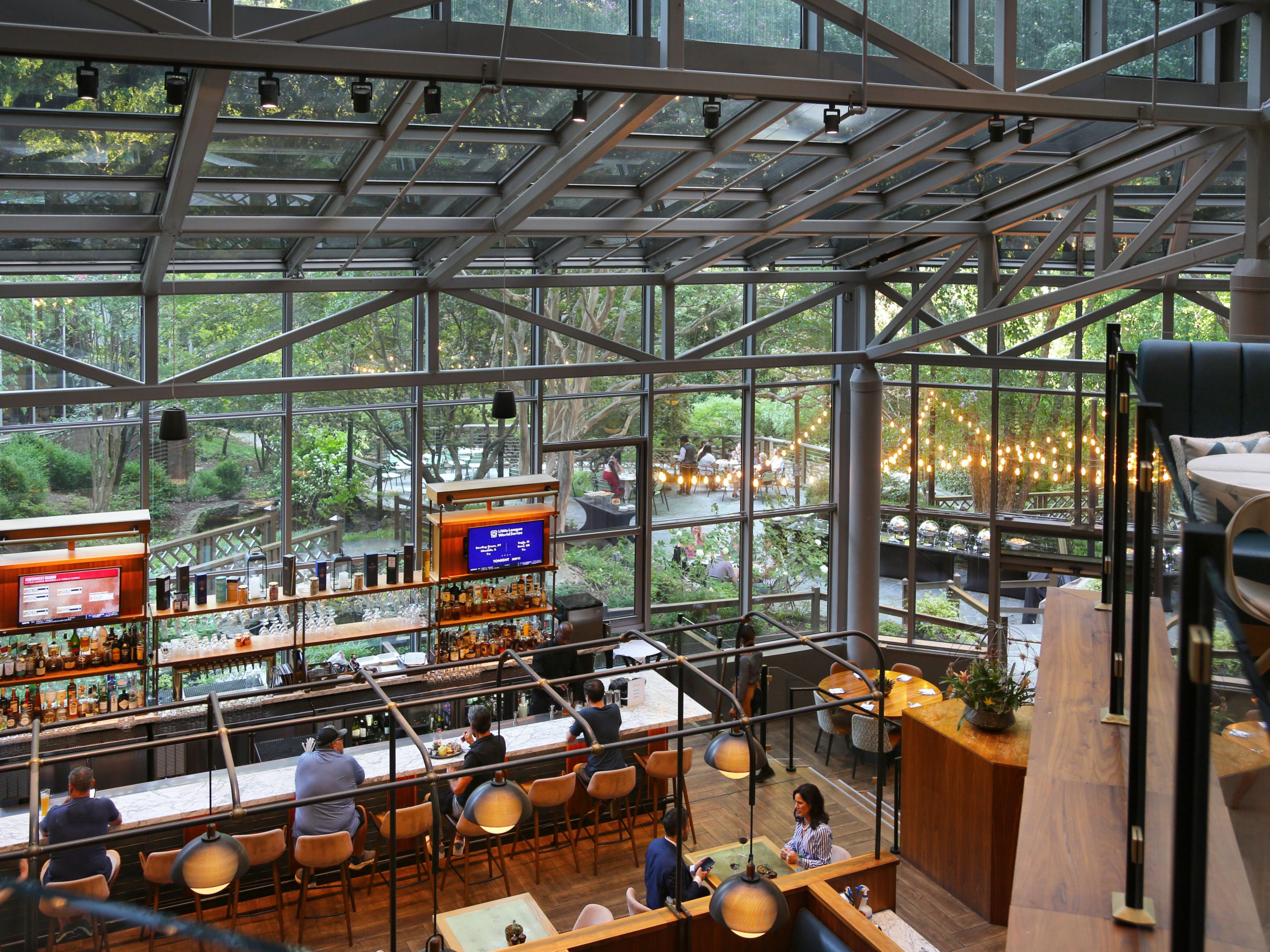 A high-angle view of a stylish indoor bar with a massive glass ceiling and walls showcasing a lush green garden. Patrons sit at the bar counters lit by warm pendant lights, while outdoor seating areas are visible through the windows, creating a seamless indoor-outdoor atmosphere.