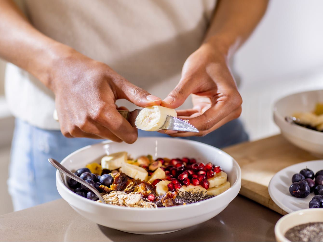 Hands slicing banana into bowl of fruit and granola