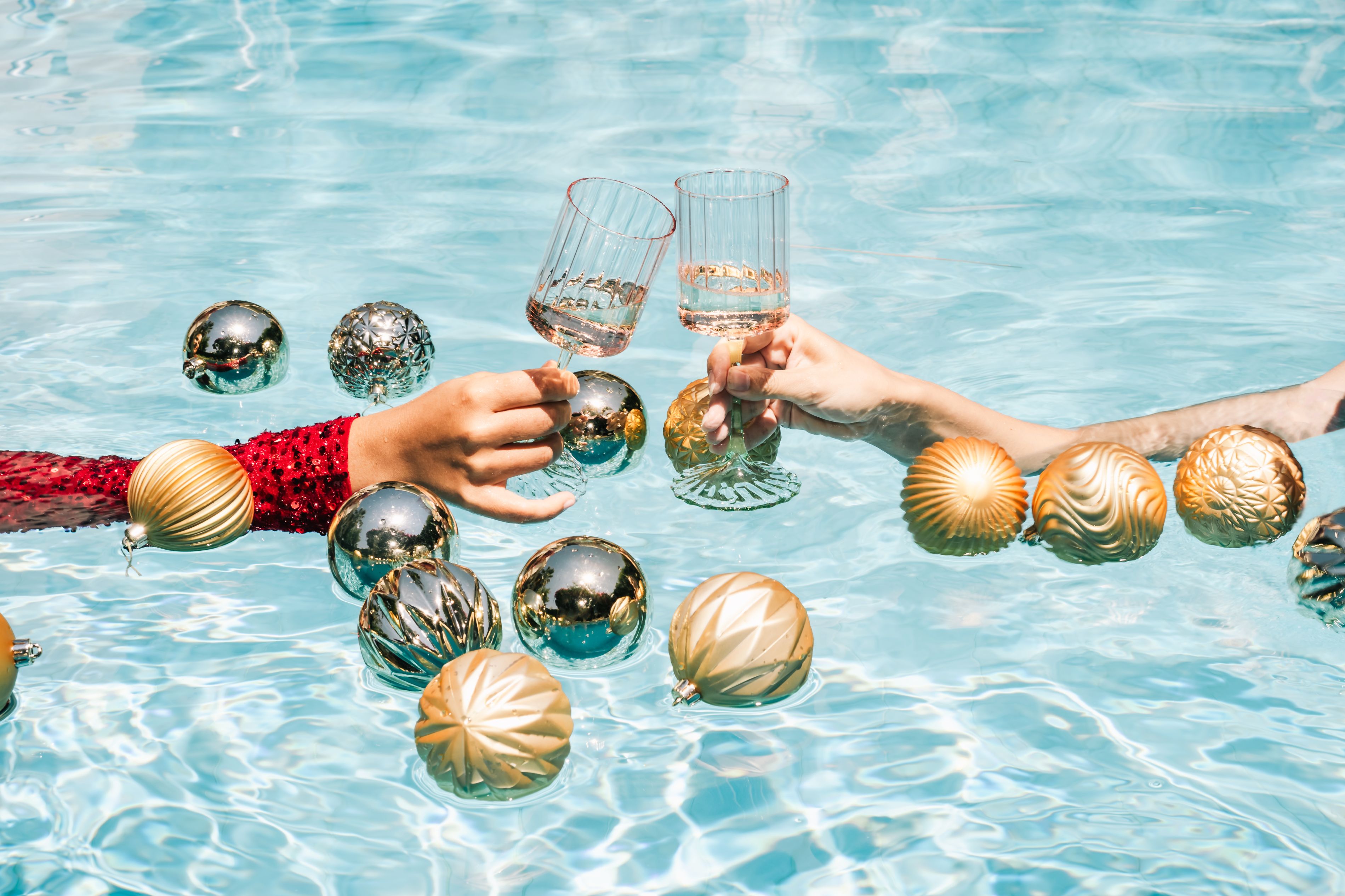Two people toast with champagne glasses in a pool surrounded by gold and silver holiday ornaments.